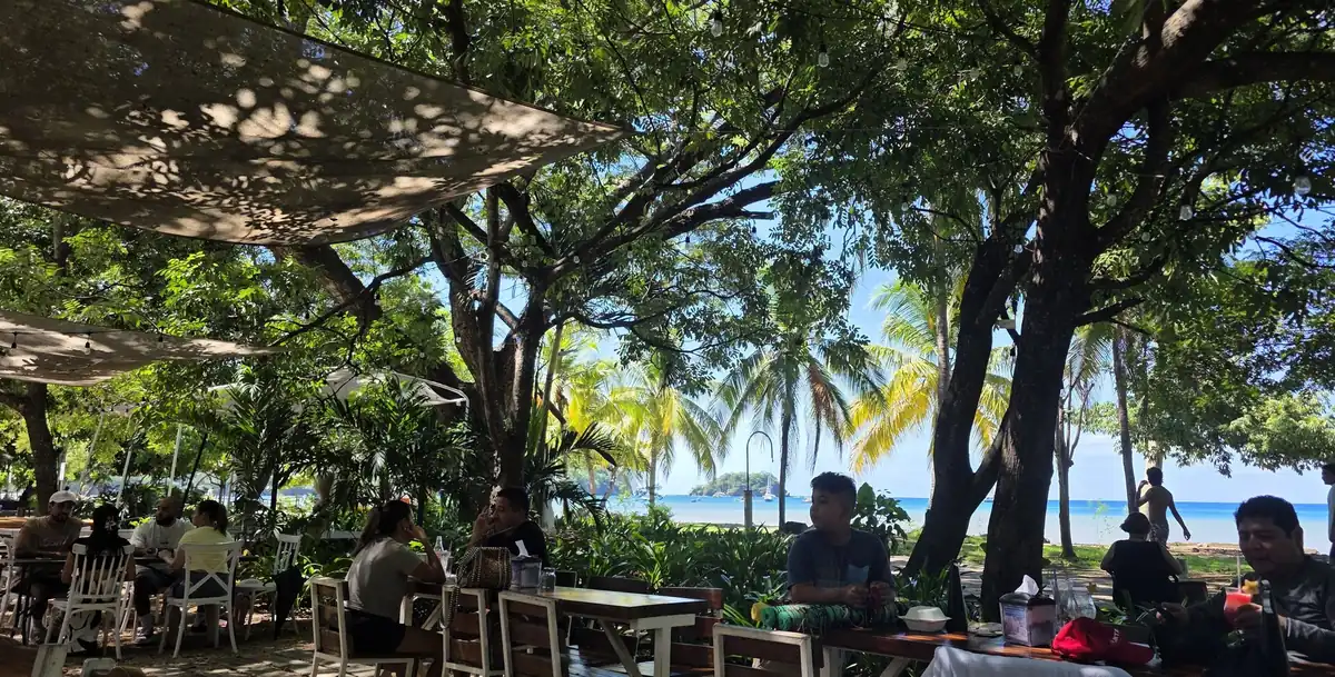 Mesas al aire libre bajo la sombra de los árboles en el Restaurante Donde Claudio y Gloria con vista a las palmeras, el mar turquesa y los botes en la bahía de Playas del Coco