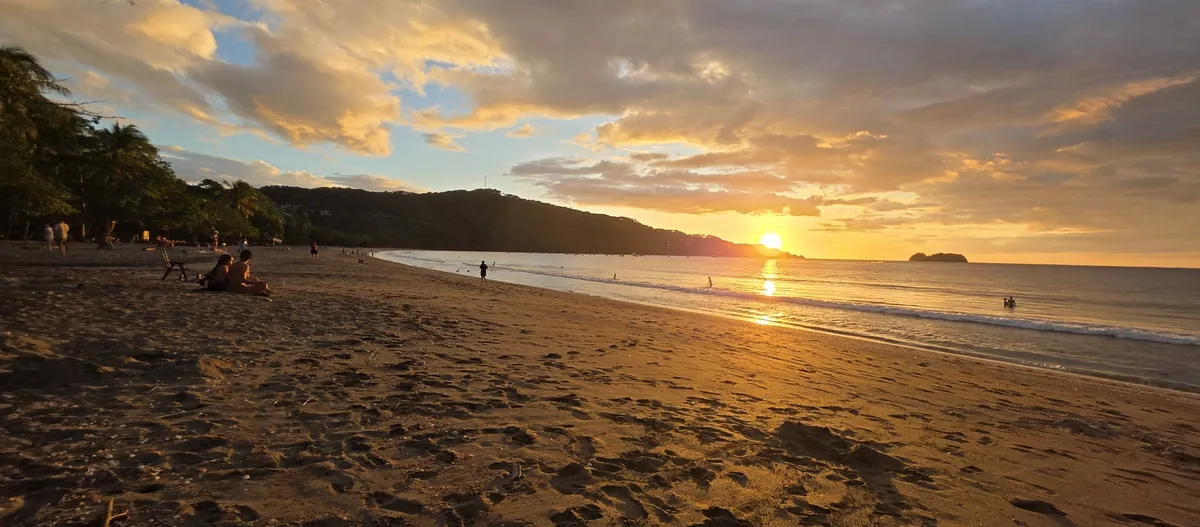 Atardecer dorado sobre la bahía tranquila de Playa Hermosa cerca de Playas del Coco, con luz ámbar reflejándose en el mar suave y personas descansando en la arena