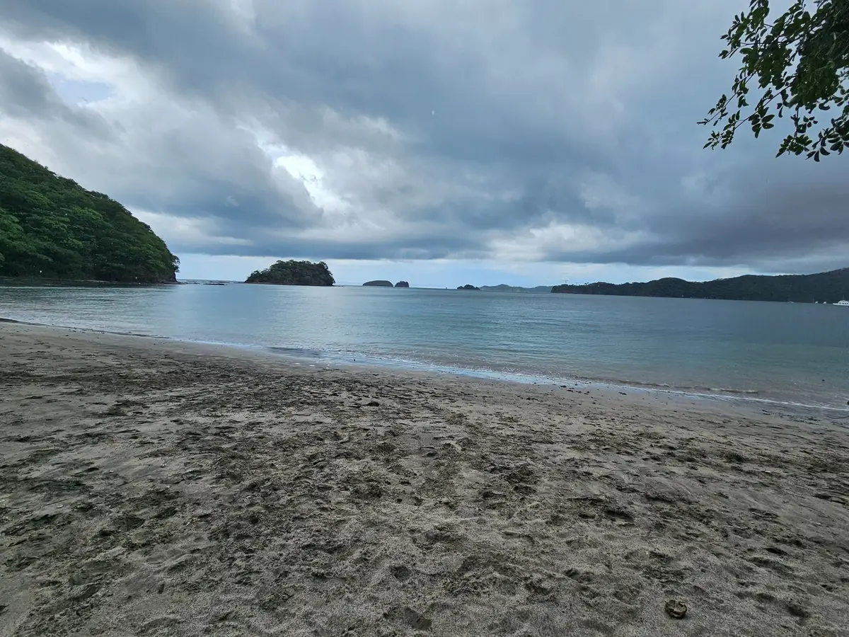 Amplia franja de arena volcánica oscura en la playa de Playas del Coco con olas suaves, colina verde e islotes rocosos al fondo de la bahía